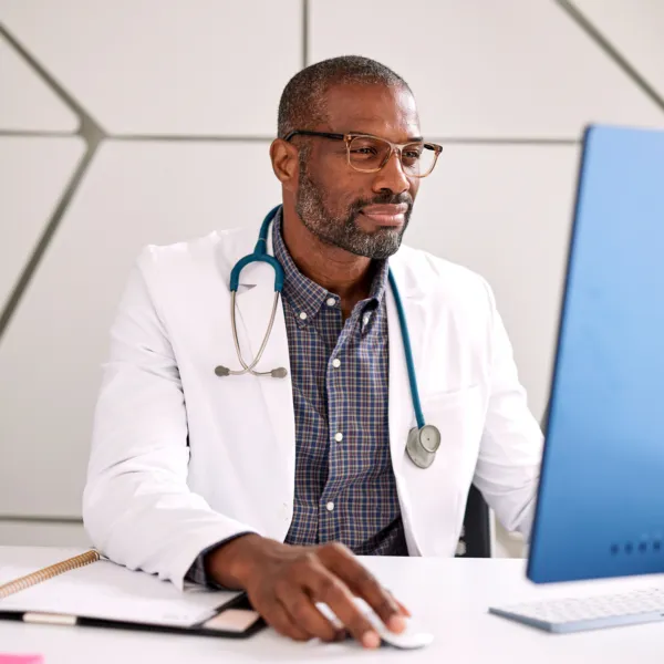 Black male doctor wearing glasses, a white lab coat, and a stethoscope sitting in front of a computer in an office