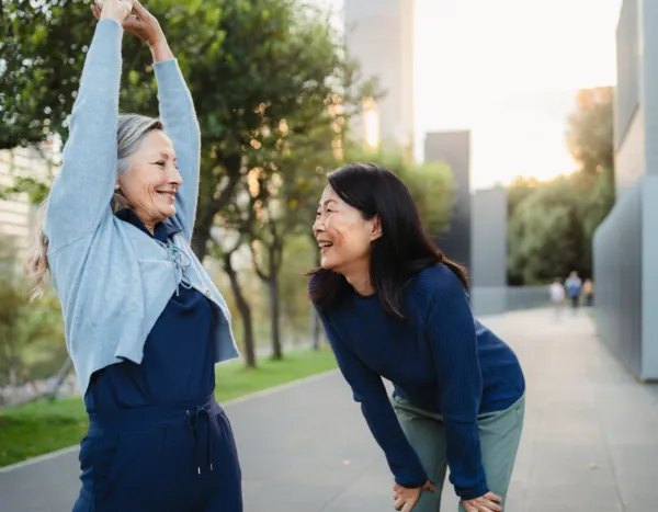 brown haired woman in gray and pink athletic clothes and blonde haired woman in blue athletic clothes stretching and laughing outside