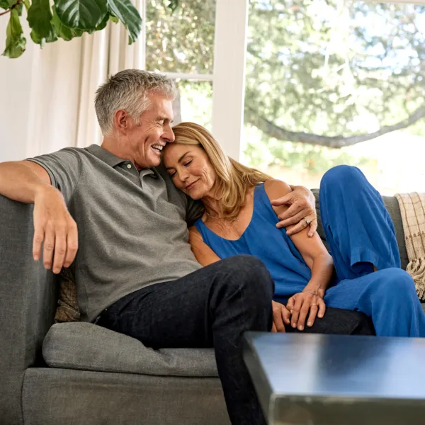 white man with short gray hair in gray top and jeans sitting on a couch in front of an open window with his arm around a blonde woman in blue jumpsuit