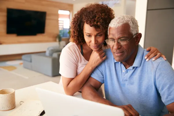 a black couple in their 50's or 60s looking at a computer. Male has white hair and female has caramel and red highlights in her dark brown hair.