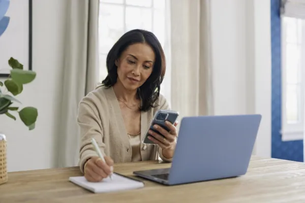 woman in a tan sweater with medium length dark hair sitting in front of a laptop at her dining room table writing on a notepad with one hand and holding a phone in the other