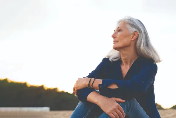 white woman with gray hair in a blue shirt sitting outside with her arms wrapped around her knees and gazing off camera