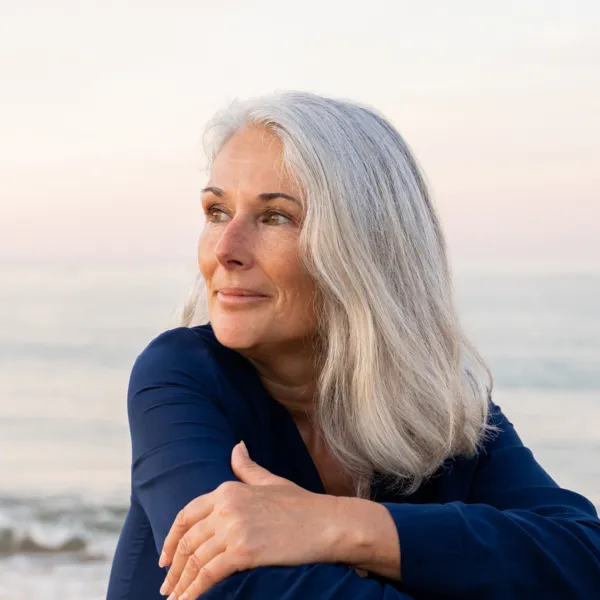 waist up portrait of a white woman with gray hair in a blue shirt on the beach looking off camera and smiling slightly