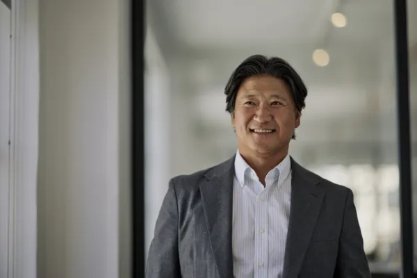 waist up view of a smiling Asian male with brown hair wearing a white button up shirt and a gray jacket