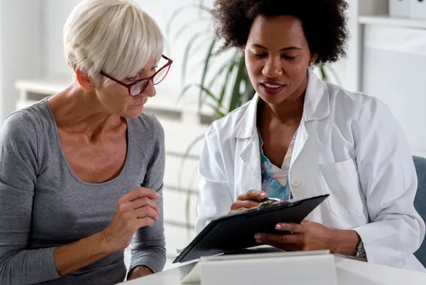 black female doctor in lab coat presenting data on a folder and talking to a white blonde female patient in a grey long sleeved shirt.