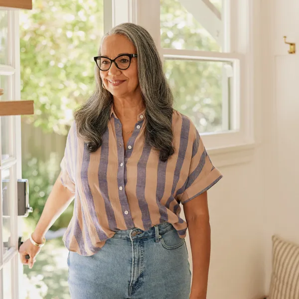 Black woman with long gray hair wearing black cat-eye glasses, a pink and blue striped shirt, and jeans smiling in front of an open door in her home