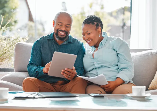 Black couple sitting at their kitchen table writing on a notepad with a cup of coffee and a tablet on the table in front of them