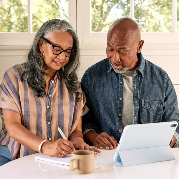 Black couple sitting at their kitchen table writing on a notepad with a cup of coffee and a tablet on the table in front of them