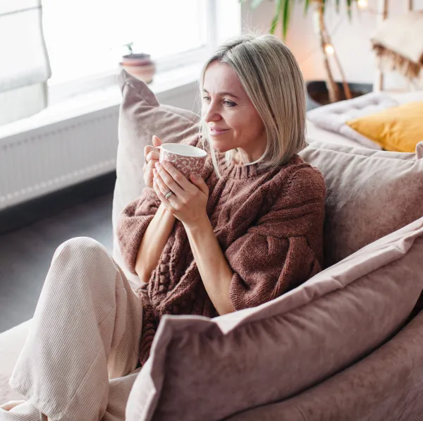 woman with brown hair wearing a tan sweater and gray pants smiling softly and sitting on a chair in her living room