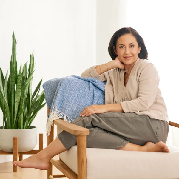 woman with brown hair wearing a tan sweater and gray pants smiling softly and sitting on a chair in her living room