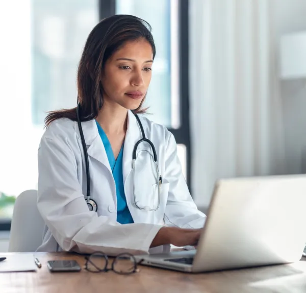 Woman sitting in front of a lap top wearing a white lab coat and stethoscope and writing on a notepad