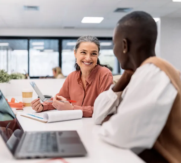 Black woman with short hair wearing a white polka dot shirt with a gray jacket sitting at a table smiling at an out of focus woman in a blue and black shirt