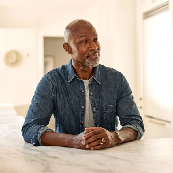 Bald Black man wearing a button up denim shirt sitting at a table with his hands folded in front of him